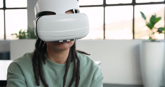 Non-binary African adult in office chair wearing white VR headset, rotating tablet, testing VR app