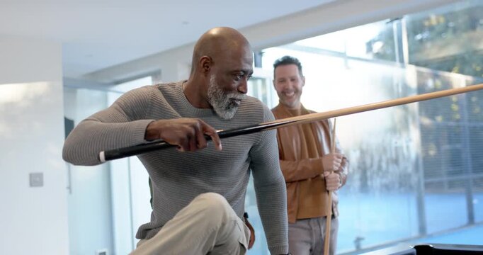 Diverse men watching mature man picking cue kneeling on pool table at home, aiming hard shot