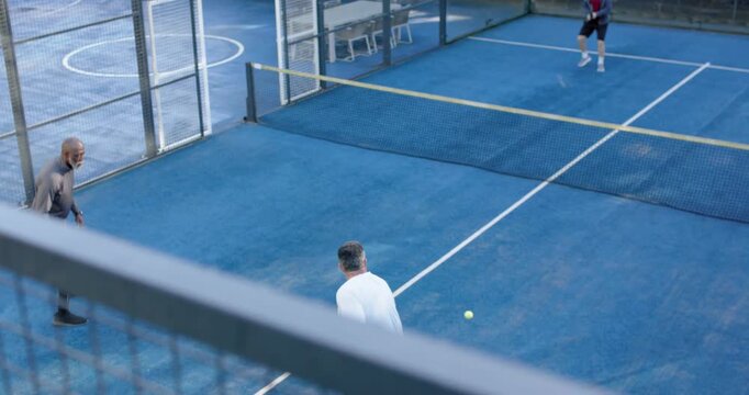 Four men playing doubles on padel court, white-shirt serving, competing returning ball with racquet