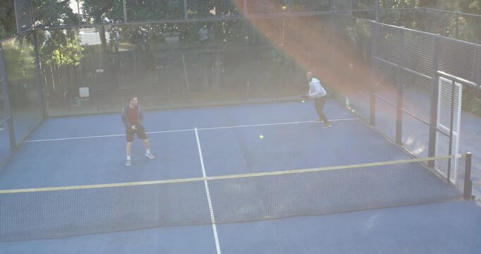 Diverse male players tracking bouncing yellow ball using paddles on blue padel court during match