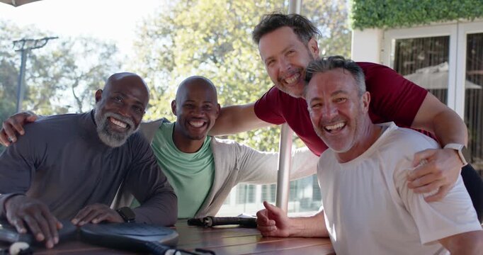 Diverse male friends posing after camera cue at shaded patio picnic table, draping arms, laughing
