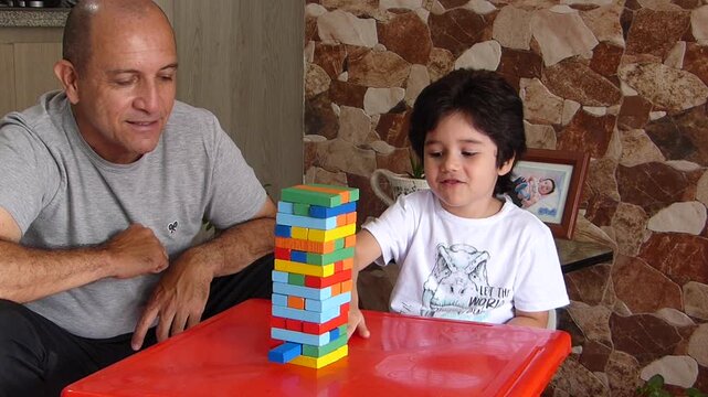 Smiling father watching his focused young son playing a colorful wooden block stacking game on a red table at home