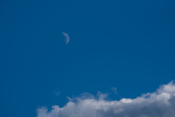 Minimalist view of a white crescent moon visible in the bright blue daytime sky above a soft cloud © Stephan