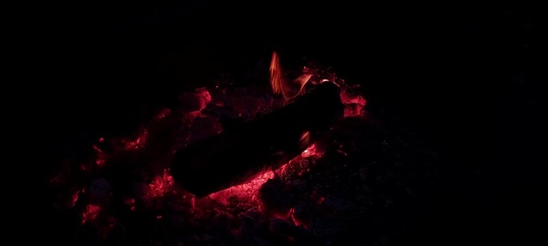 A burning log and glowing embers in a dark cinematic style in red and black colors representing a warm campfire at night with copy space