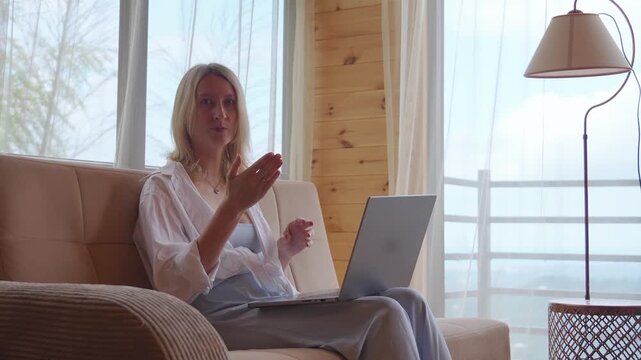 Confident female manager sitting on a sofa with a laptop, actively gesturing and speaking during an online business meeting, webinar, or remote job interview from her cozy home office