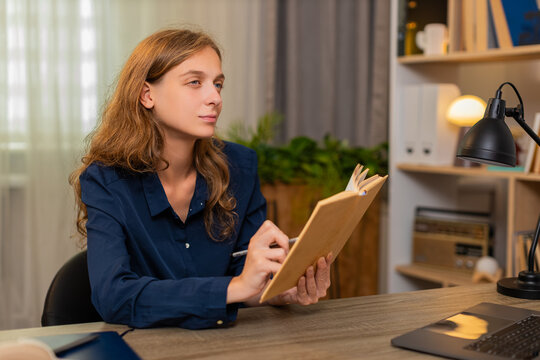 Young woman on home table reads study book underlining key line and learning with calm focus. Adult girl on home table reviews notes and continues learning with pen and laptop for calm online study.