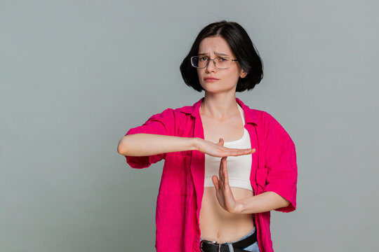 Young woman forms timeout symbol with her hands, looking serious to clearly indicate a request for a pause or break. Caucasian girl expresses the need to stop or slow down, isolated on gray background
