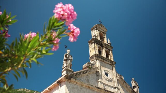 Historic stone church framed by blooming oleander flowers in Prcanj on the Bay of Kotor. Mediterranean architecture under a clear summer sky