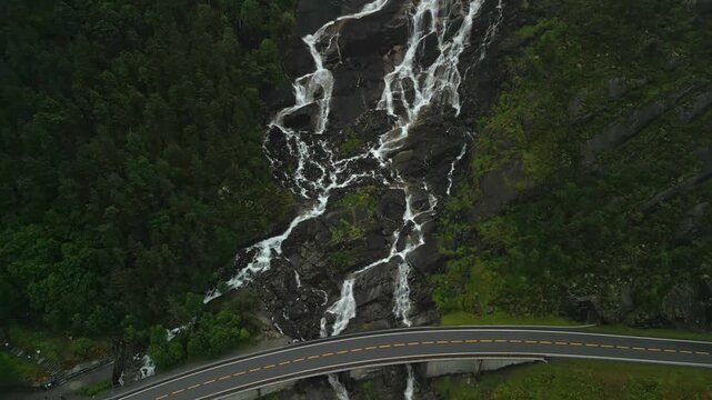 Water cascades down rocky cliffs beside a winding road, showcasing the stunning Langfoss waterfall in a lush green landscape with misty mountains in the background