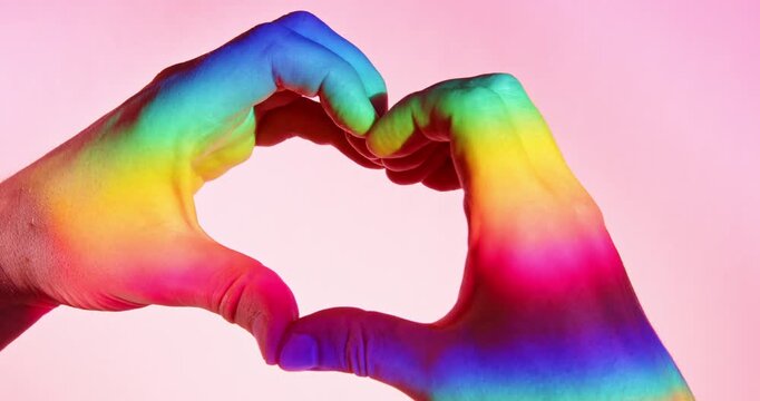 Close-up studio video of two hands forming a heart shape illuminated by rainbow-colored light against a pink background in slow motion. Symbolic LGBTQ+ Pride concept representing love and diversity