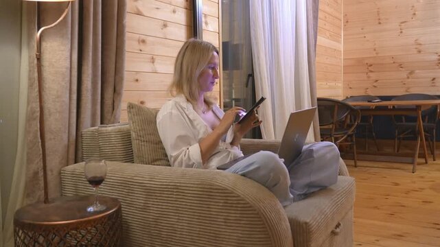 Young woman sitting comfortably in an armchair, scrolling through her smartphone while a laptop rests on her lap, enjoying a relaxing evening in a cozy, wood-paneled living room