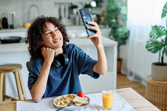 A smiling teen sits at a kitchen table taking a selfie with a smartphone while wearing headphones. A plate with scrambled eggs and toast and a glass of orange juice sit in front of them, conveying a r