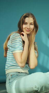 Vertical video of young happy blonde woman with long healthy hair in striped top and jeans sitting on the floor in studio on blue background