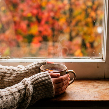 A cozy cup of hot tea held by a person in a warm sweater, with a blurred background of colorful autumn leaves outside the window