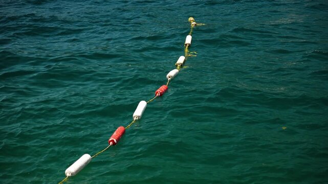 Red and white buoys connected by a rope float across clear turquoise sea water. The floating barrier marks a designated swimming area along the coast