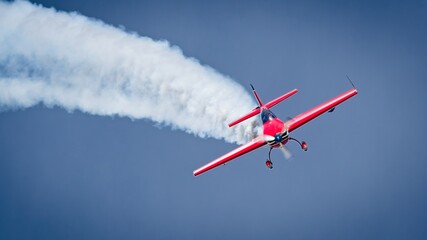 Red aerobatic stunt plane diving with a thick white smoke trail against a clear blue sky. © César
