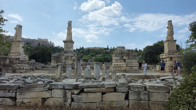 Ancient Agora, Athens, Greece - 8 July 2026. Odeon of Agrippa ruins with monumental Triton figures, stone remains and visitors, with the Acropolis in the distance.