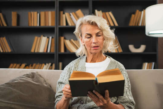 Comfort relaxed senior old elderly woman reading book at home. Mature woman resting enjoy calm day sitting on sofa at home alone. Adult lady spending free time keeping book smiling. Pension concept