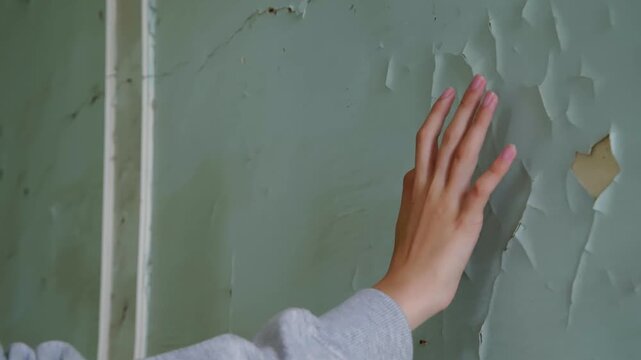Young person hand touching worn plaster wall with peeling paint, revealing texture of aged surface in green toned interior space.
