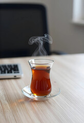 Fototapeta premium Steaming turkish tea in a traditional glass cup on a wooden desk with a laptop keyboard in the background, a laptop keyboard is visible in the blurred background, a work or study environment