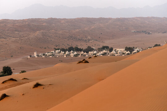 View of a desert camp with white tents nestled among vast orange sand dunes under a clear sky in Bidiyah, Ash Sharqiyah North G, Oman.