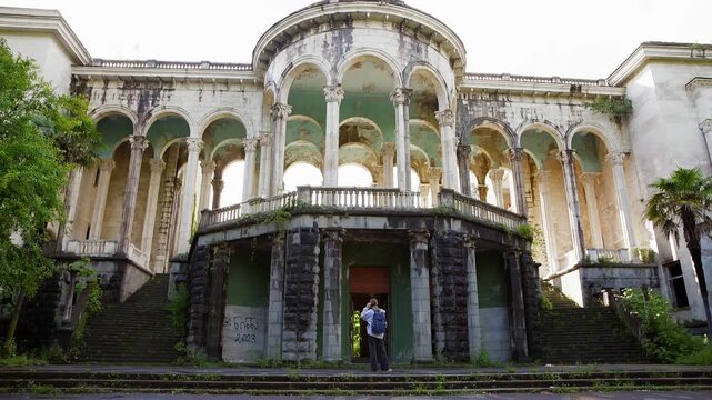 Solitary visitor explores decaying neoclassical palace featuring towering colonnades, weathered stone arches, moss covered steps, lush vegetation reclaiming ornate architecture.