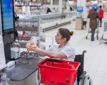 A Caucasian woman in a wheelchair pays for groceries at a self-checkout counter in a supermarket.