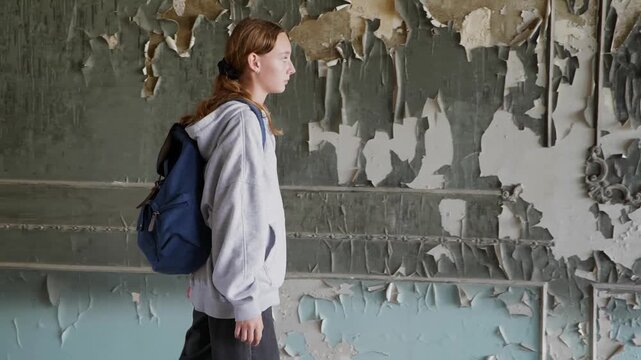 Woman in casual clothing walks by heavily damaged, peeling paint on walls inside dilapidated interior space, depicting decay, abandonment, urban exploration.