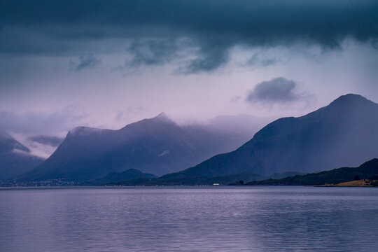 View of the moody mountains rising above the calm waters of the fjord under a dark, overcast sky in Sortland, Nordland, Norway.