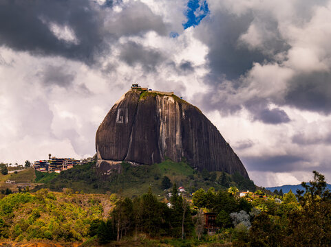 View of La Piedra monolith rising above lush green hills under a dramatic cloudy sky in La Piedra, Antioquia, Colombia.