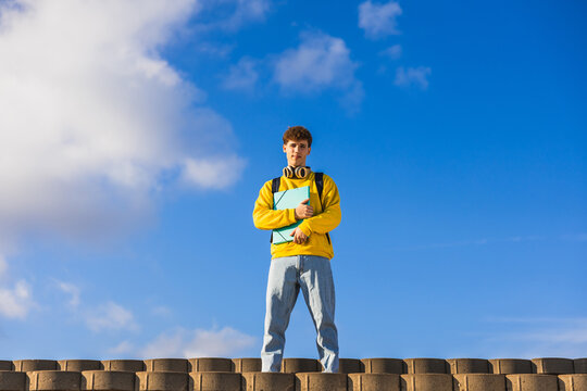 College student holding book and standing on concrete steps with blue sky background