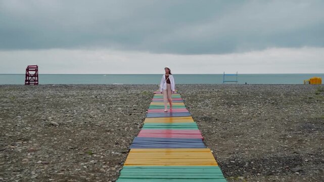Woman strolls along vibrant rainbow striped wooden pathway on rocky beach with calm sea horizon, overcast sky, lifeguard tower visible in background.