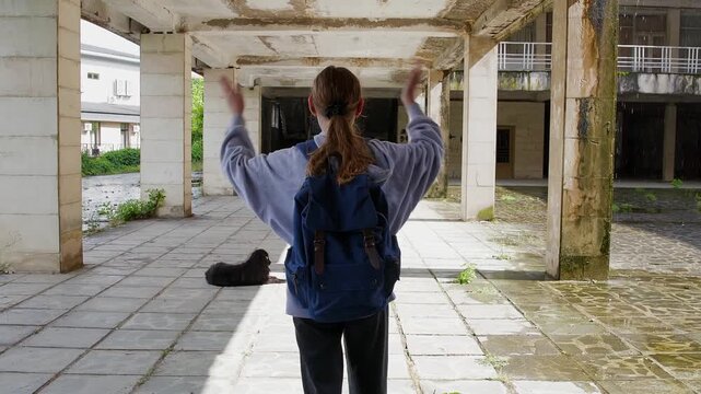 Woman in casual attire walks through deteriorating covered passageway while black dog lies on paved ground in foreground, sunlight streaming across overgrown abandoned structure.