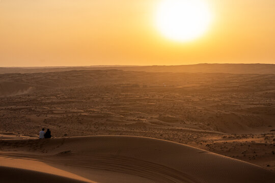 View of two people sitting on a sand dune in the desert during golden hour with the sun setting over the vast landscape in Bidiyah, Ash Sharqiyah North G, Oman.
