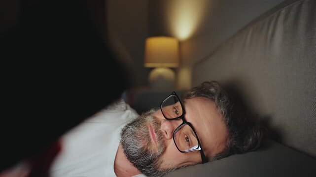 A man in eyeglasses lying in bed using smartphone late at night