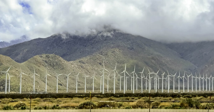 View of wind turbines in the desert with mountains in the background under a cloudy sky in Palm Desert, California, United States.