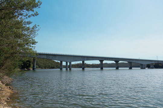 Concrete bridge spanning a calm body of water, surrounded by trees and captured under a clear blue sky, illustrating transportation infrastructure, civil engineering and landscape.