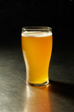 Glass of golden beer with a creamy white foam head, photographed on a dark metal surface against a black background. Dramatic studio lighting highlights the beer&rsquo;s amber color, glass texture