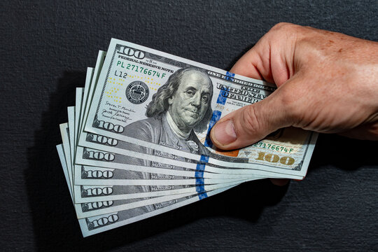 Closeup of senior or mature hand holding fan of crisp US one hundred dollar banknotes against dark textured background