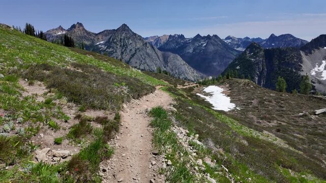 First-person point of view moving forward along a dirt mountain path with snowfield patches and dramatic mountain range in the background on a sunny day in the North Cascades, Washington