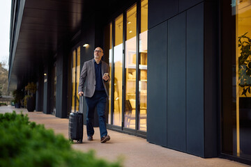 Fototapeta premium Mature businessman with smart phone and travel bag walking on street.