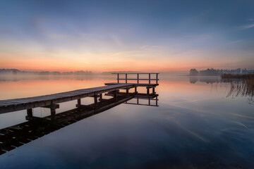Fototapeta premium Wooden jetty on a calm misty lake at sunrise with pastel sky reflection, long exposure peaceful morning landscape
