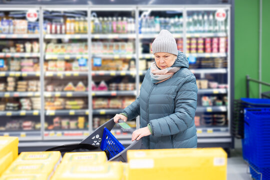 Elderly female customer wearing scarf choosing groceries while standing with trolley in contemporary retail store interior.