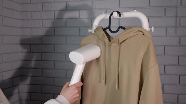 Woman's hand steaming beige hoodie with garment steamer. Close-up of a person steaming a beige hoodie with a portable iron steamer against a grey brick wall, performing household chores