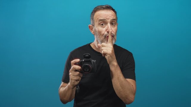 Man middle aged hispanic holds camera and presses finger to lips for silence against teal studio backdrop; secretive hush calm.