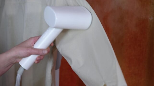 Handheld garment steamer removing wrinkles from shirt. Housewife's hand holding a portable vertical steaming iron to smooth out wrinkles on a hanging shirt sleeve, providing modern garment care