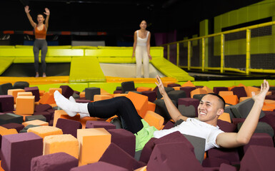 Fototapeta premium Cheerful young man enjoying soft landing in foam pit at trampoline park, with friends jumping around