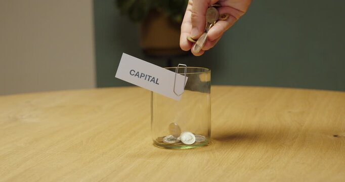 Hand placing a few coins into a jar labeled &ldquo;Capital,&rdquo; representing small capital, limited funds, low investment budget and financial constraints.