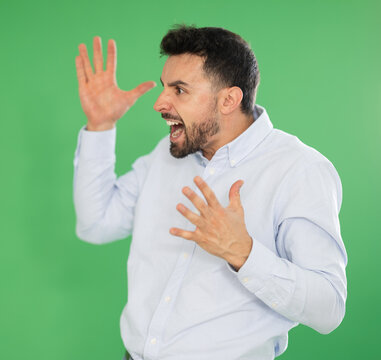 Adult man stands against green screen with angry expression and shouting or gesturing aggressively