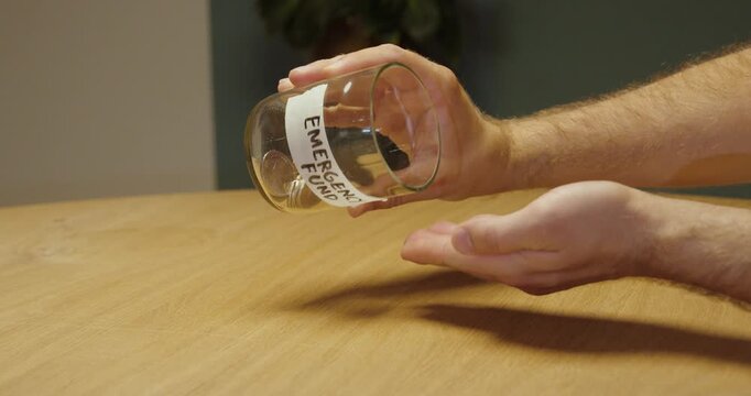 Person emptying the last coins from a glass jar labeled &ldquo;Emergency Fund,&rdquo; representing depleted savings, unexpected expenses, financial hardship and lack of emergency money.